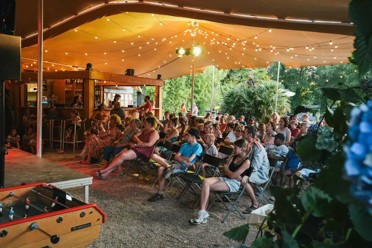 An audience seated under a stretch tent strung with festoon lights, watching a show next to a bar